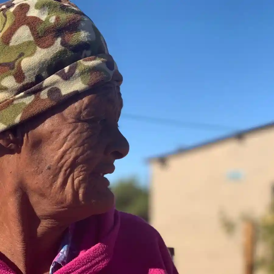 Elderly woman in a camouflage hat and pink fleece standing outdoors in a rural Northern Cape community under a clear blue sky